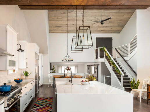 Open-concept kitchen and dining area with white cabinetry, white island with waterfall edge, wood ceiling with pendant lights, and stairs visible to the right.