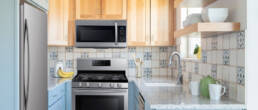A remodeled kitchen featuring light wood cabinets, blue lower cabinetry, granite countertops, stainless steel appliances, patterned tile backsplash, open wooden shelving, and a connected work area.