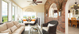 A bright sunlit living room with exposed brick arches, cream-colored upholstered seating, black wicker furniture, skylights, ceiling fan, and expansive windows overlooking countryside landscape.