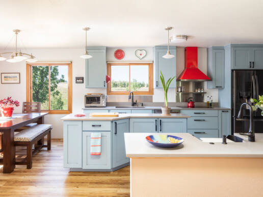 A modern kitchen and dining area remodel featuring light blue cabinetry, wooden-framed windows, a red range hood, stainless steel appliances, white countertops, and wood flooring throughout an open-concept space.