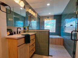 A modern primary bathroom featuring a wood vanity with quartz countertop, walk-in shower with teal subway tile, soaking tub, pendant lighting, and large mirror.