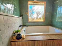 A modern bathroom featuring a white soaking tub with wood surround, green tile walls with white subway tile wainscoting, black fixtures, a window with wood frame overlooking bare trees, and decorative accessories on the tub ledge.